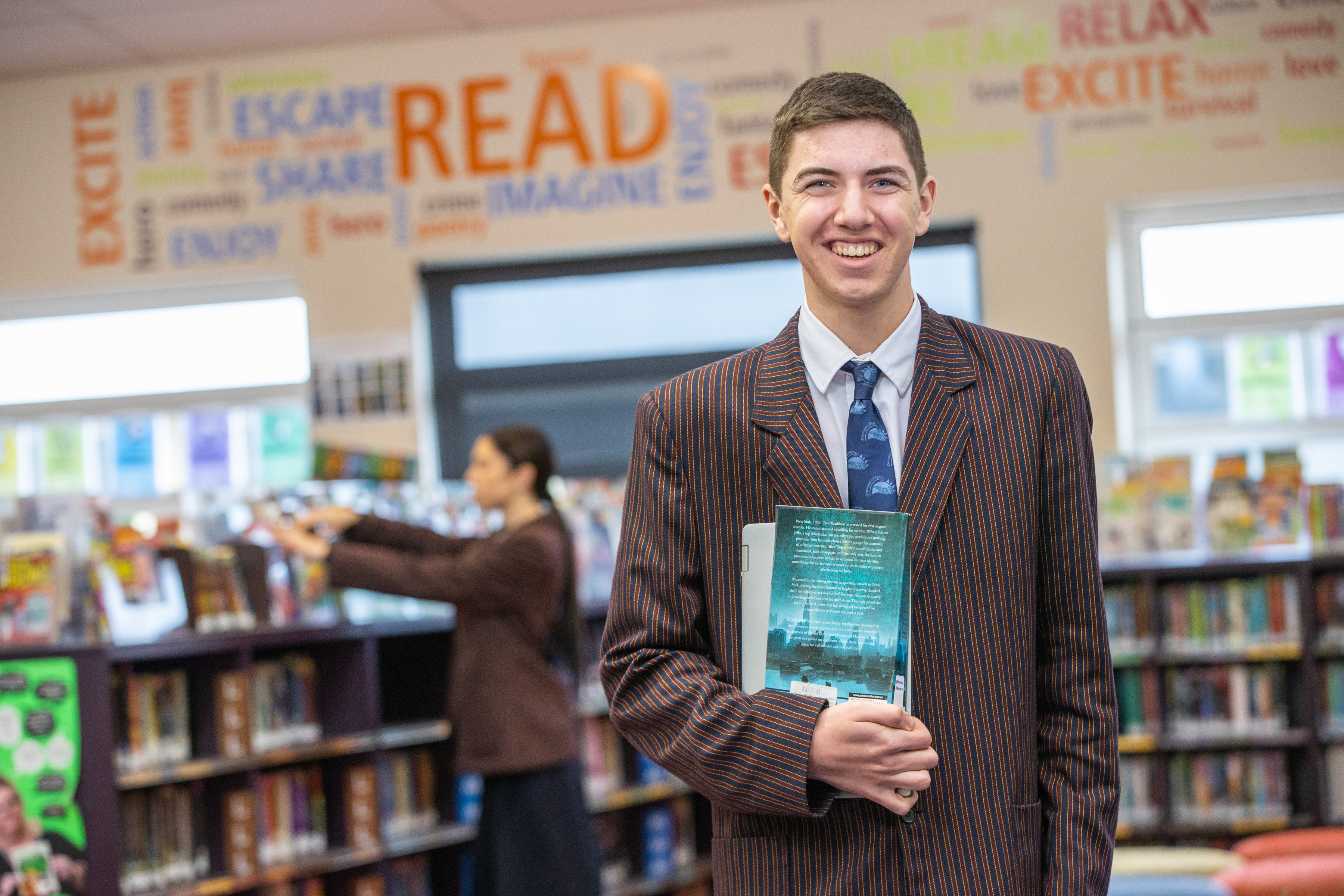 Student standing in library, holding a book, looking at the camera
