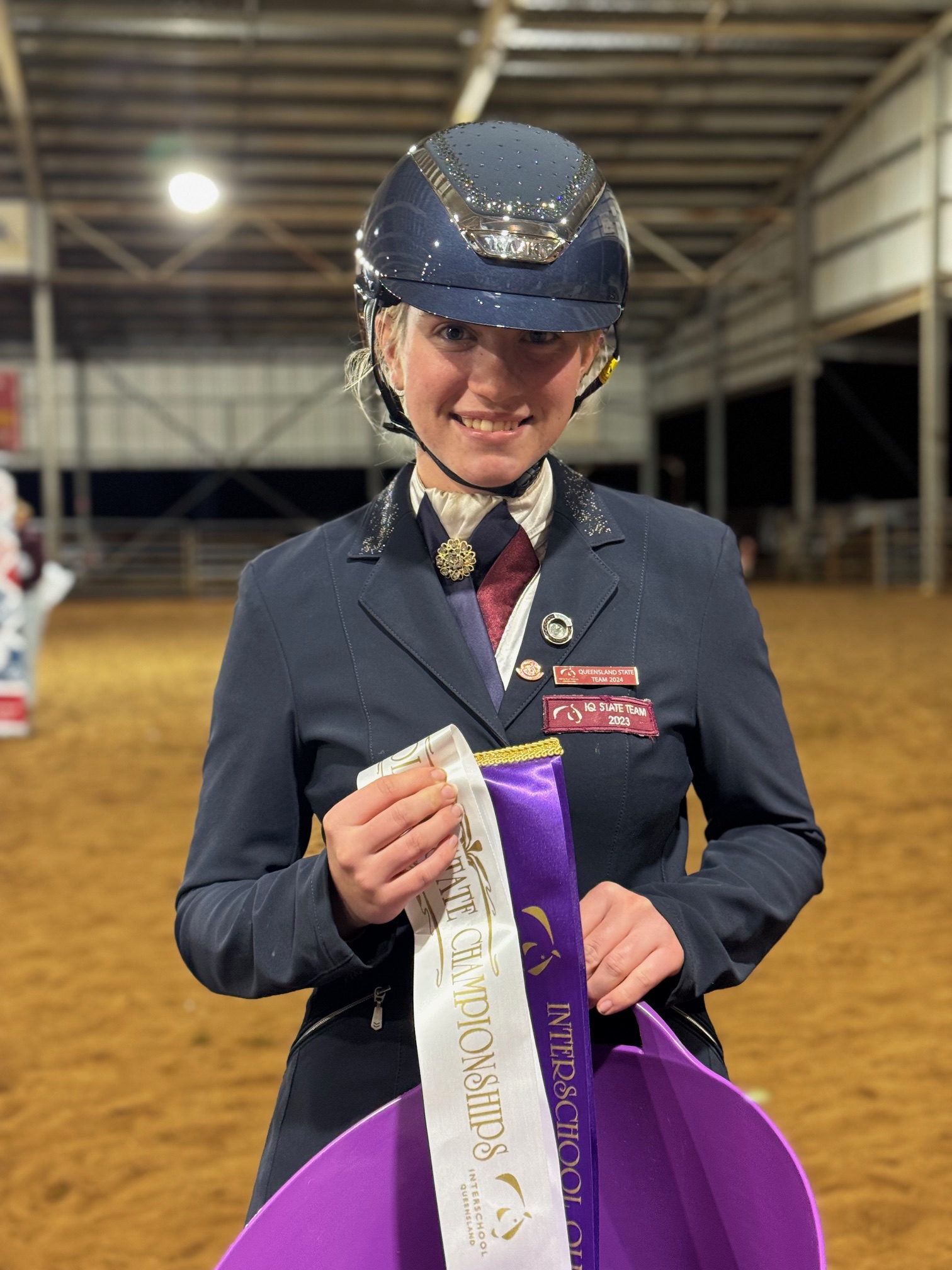 Student rider in equestrian uniform holding a championship ribbon at an indoor arena