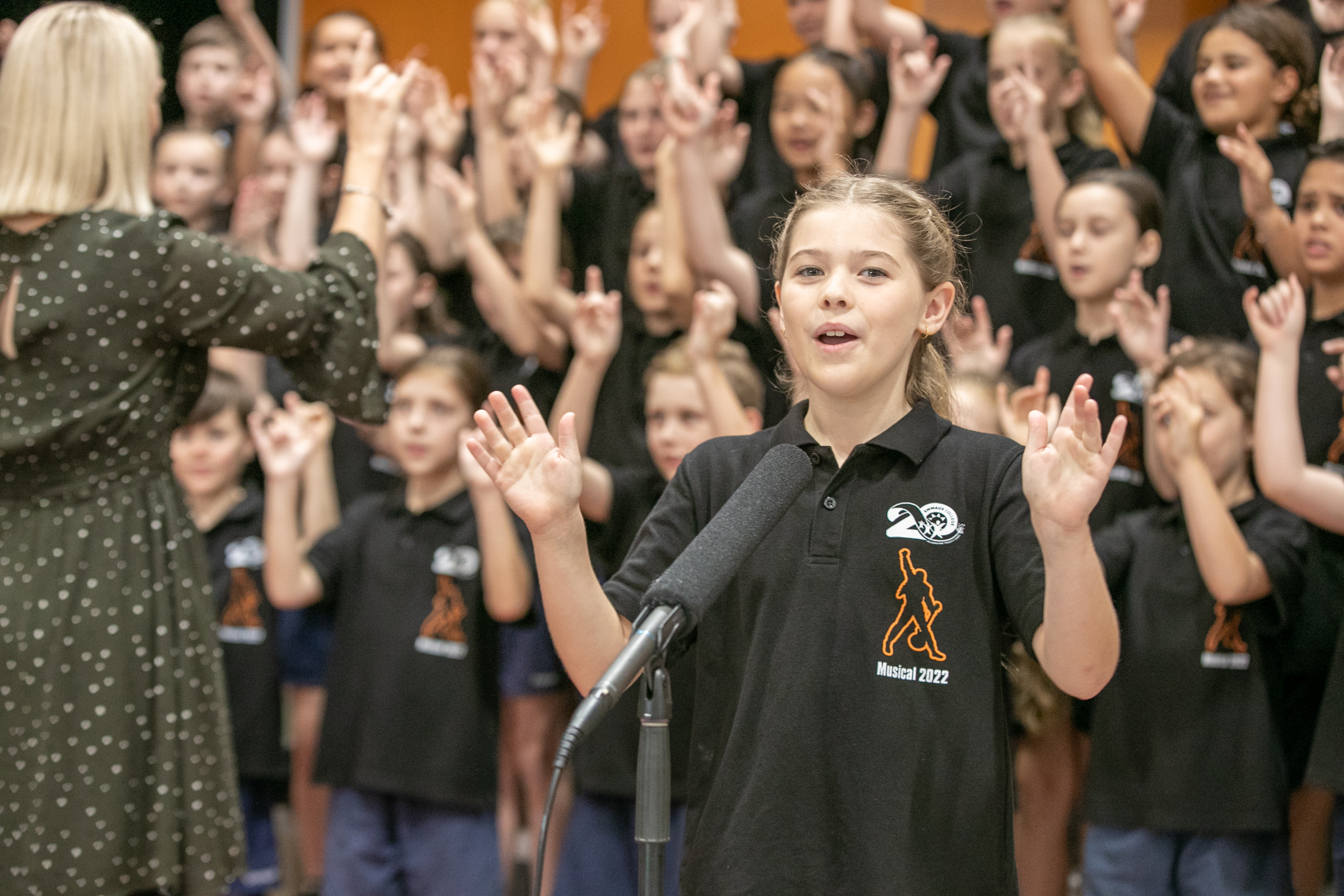 Students singing in a choir with hands in the air, being led by a Choir master