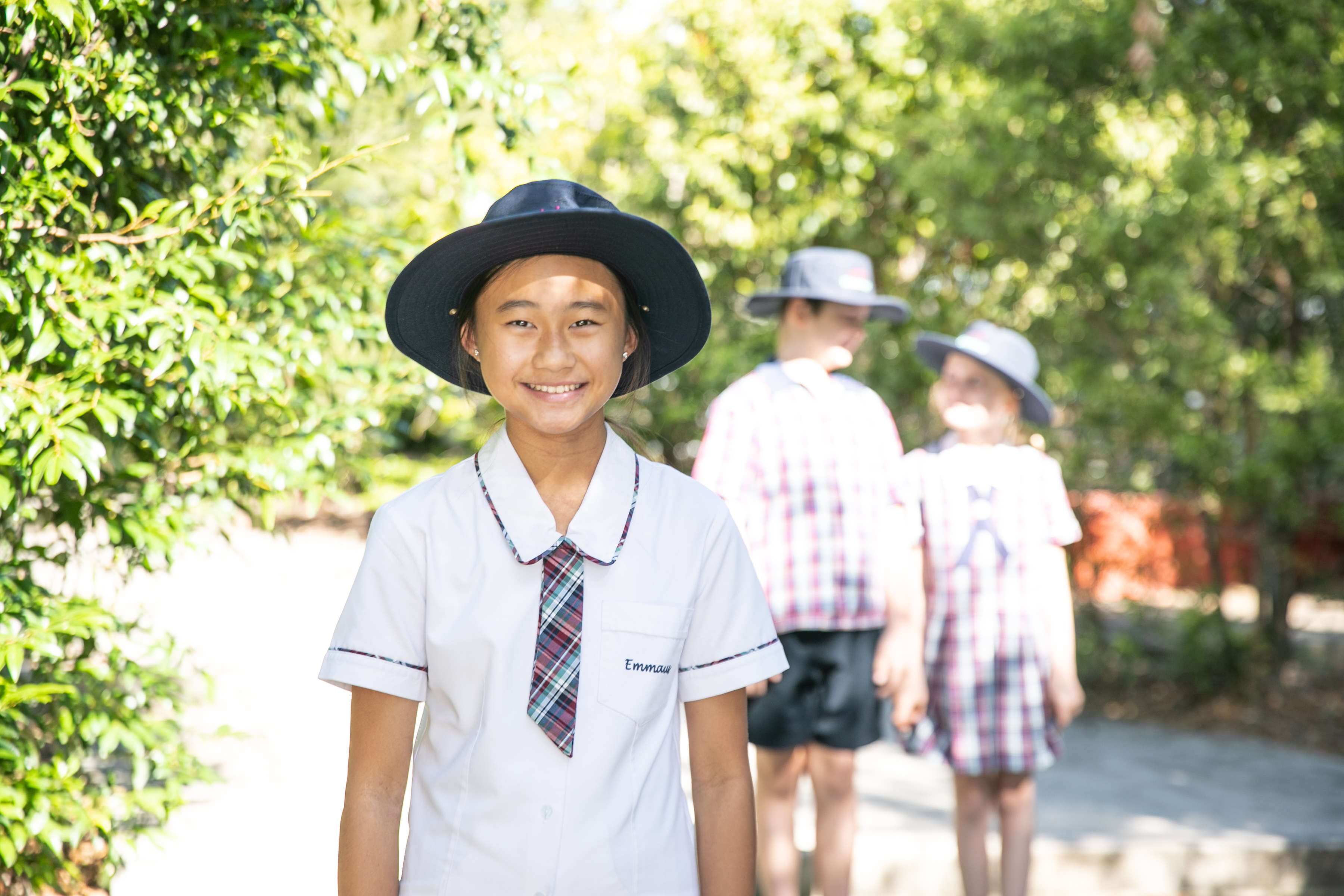 Student in uniform standing outdoors with classmates walking along a shaded school pathway