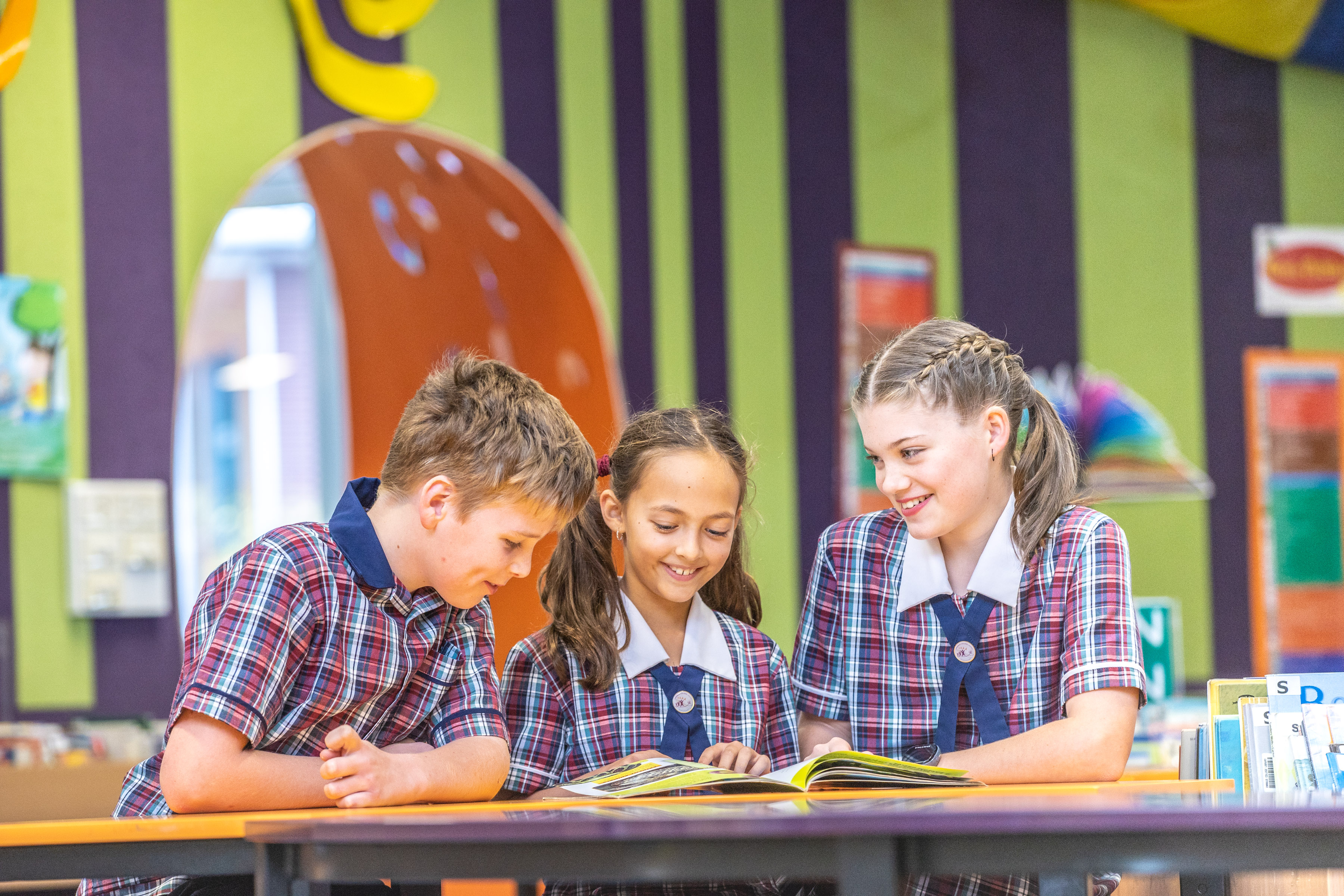 Primary school students in uniform working together at a table reading a book in a colourful library learning space
