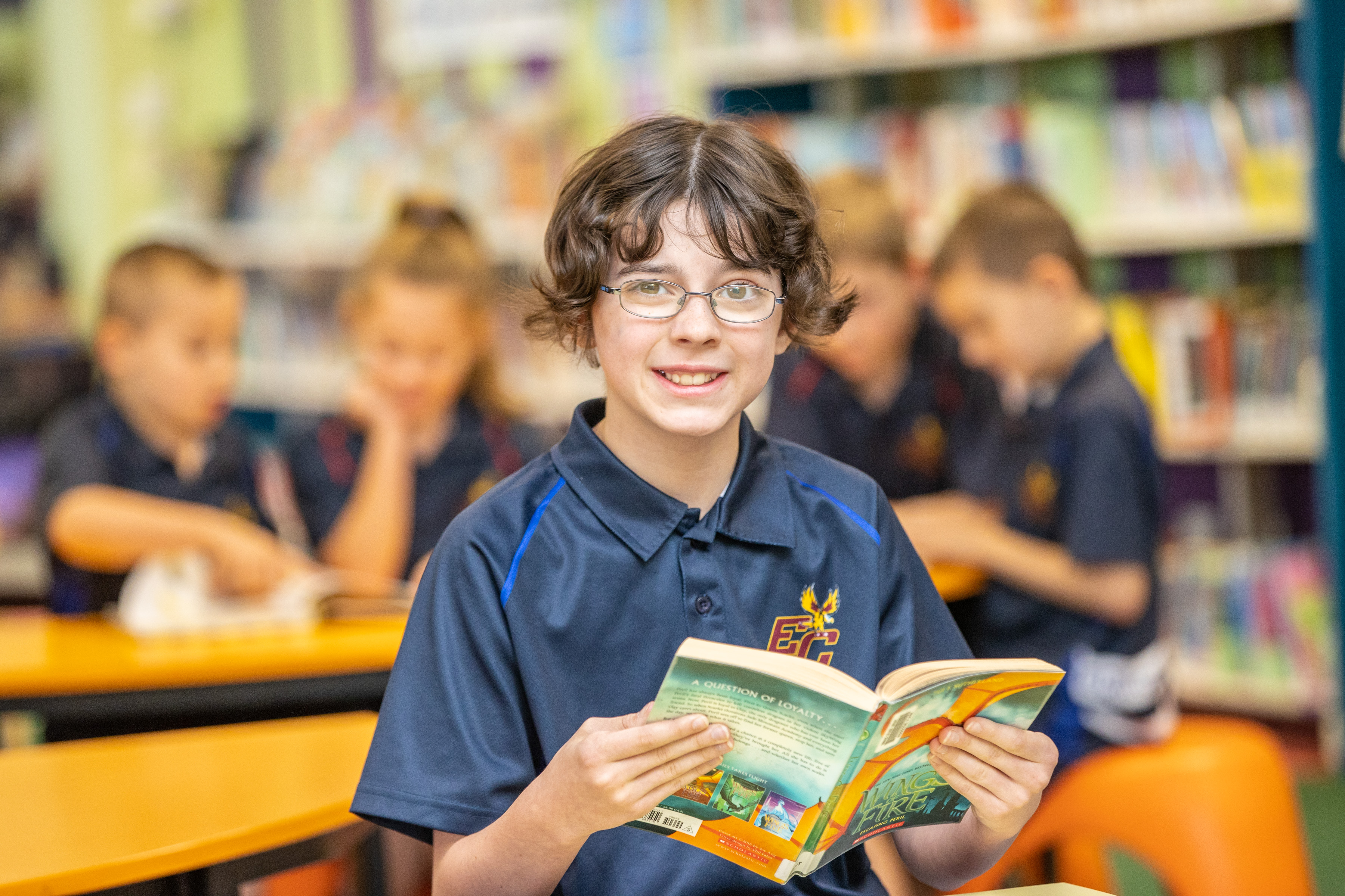 Student reading a book in a school library with classmates engaged in reading in the background