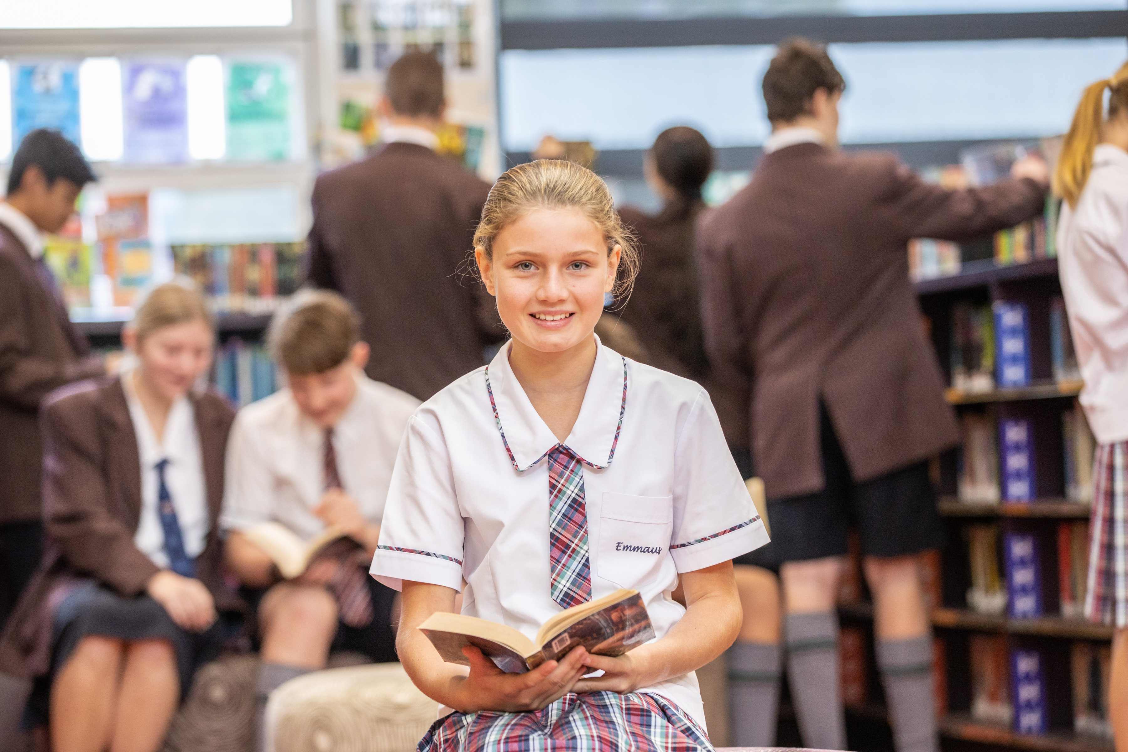 Student in school uniform seated in a library holding a book with other students reading and browsing books in the background